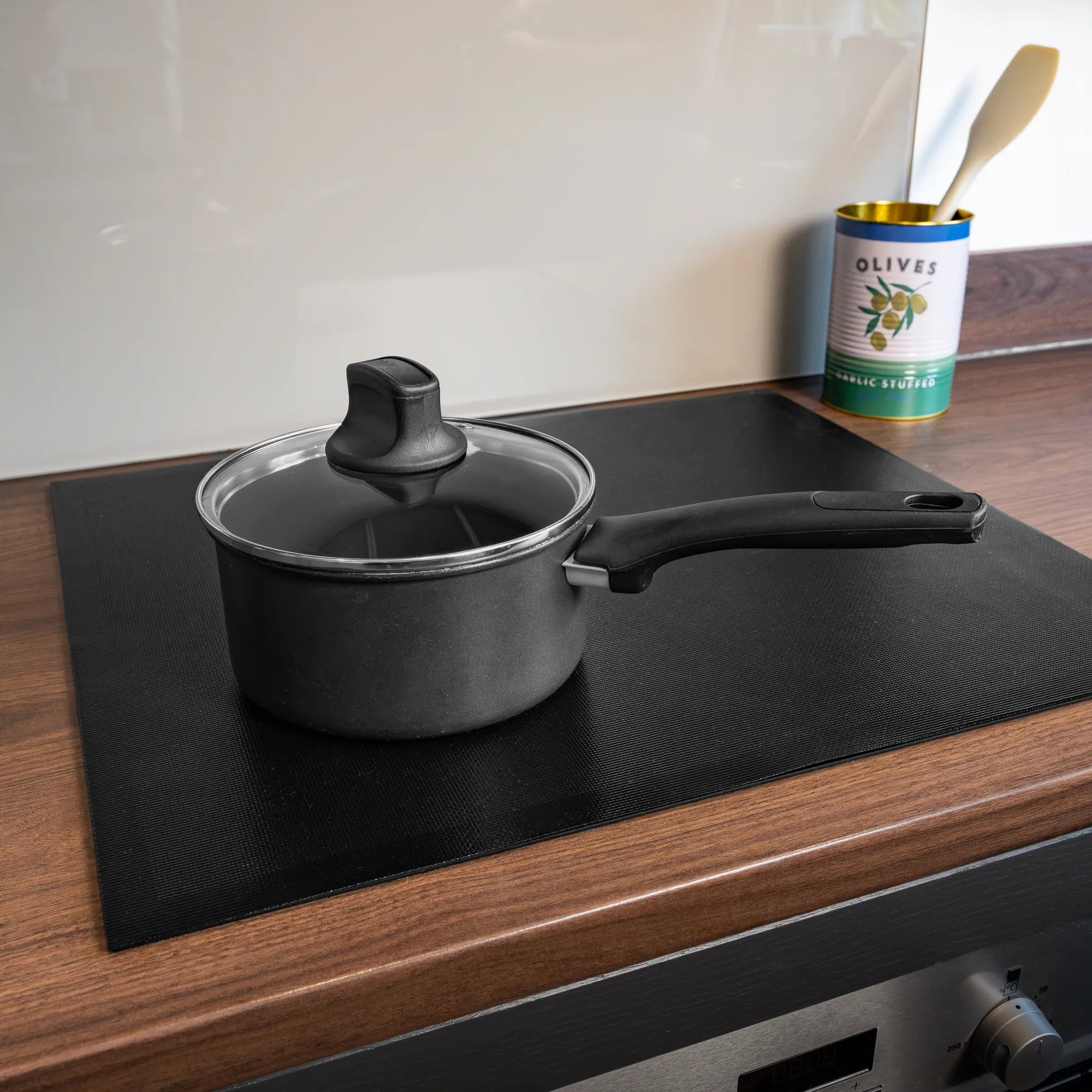 A black non-stick saucepan with a glass lid sits on a Bake-O-Glide induction hob protector on a wooden kitchen countertop. An empty can of olives and a wooden spoon are visible in the background against a light-colored backsplash.