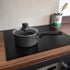 A black non-stick saucepan with a glass lid sits on a Bake-O-Glide induction hob protector on a wooden kitchen countertop. An empty can of olives and a wooden spoon are visible in the background against a light-colored backsplash.